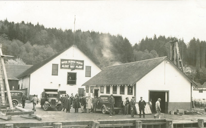 Three trucks and several people on the wharf at the Alert Bay Cannery. A sign on the building reads "British Columbia Packers Alert Bay Plant."