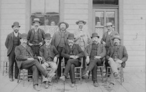 Photo de groupe avec cinq hommes assis et cinq autres hommes se tenant derrière eux. Quatre personnes regardent par la fenêtre du bâtiment qui se trouve derrière les hommes.