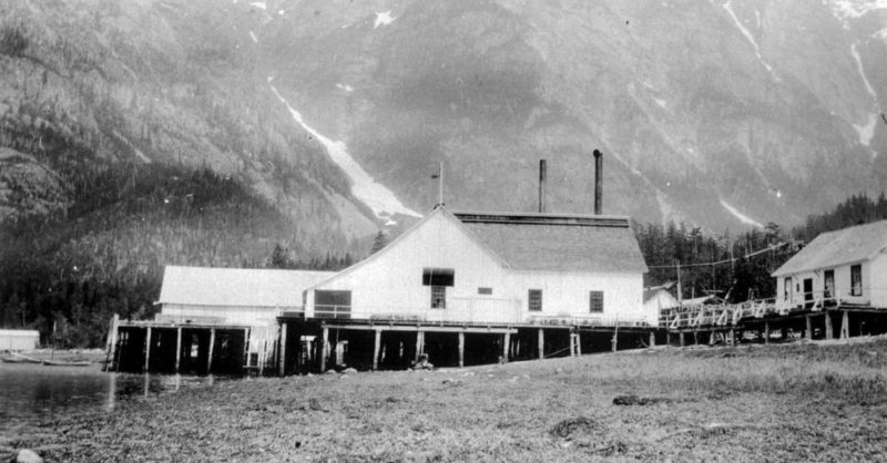 Les bâtiments de la conserverie et le quai sur piliers avec des montagnes en arrière-plan.