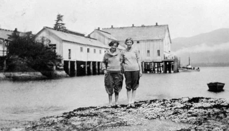 Two women stand together on a rocky point with the cannery buildings directly behind them across the water.