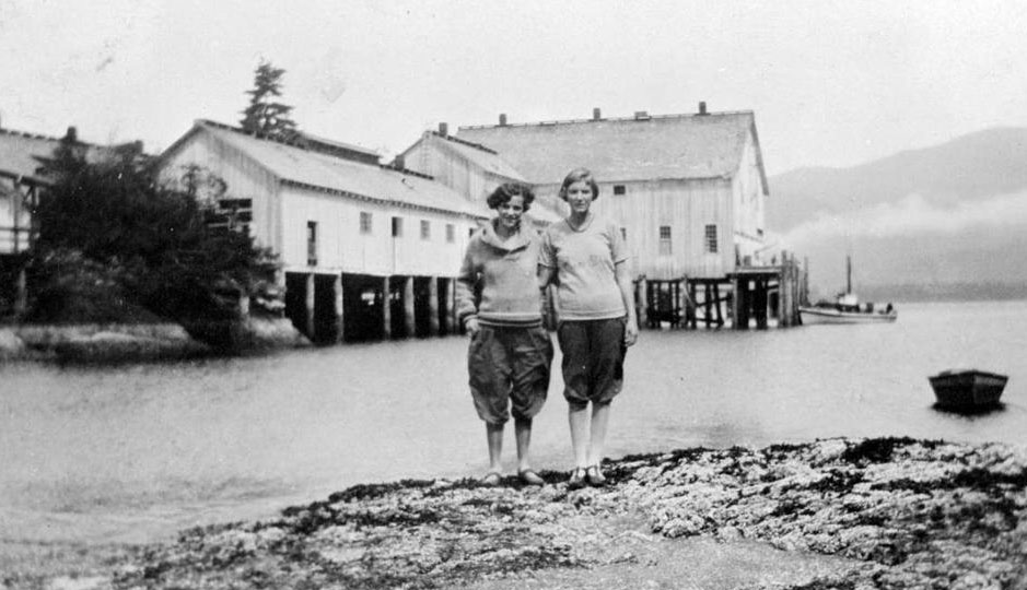 Two women stand together on a rocky point with the cannery buildings directly behind them across the water.