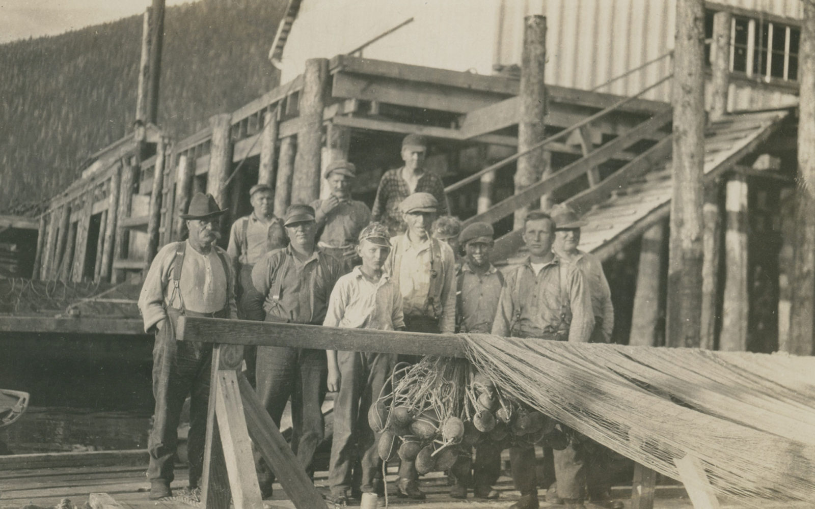 A group of men and boys stand behind a net rack on the wharf of the cannery. A net is hanging on the net rack.