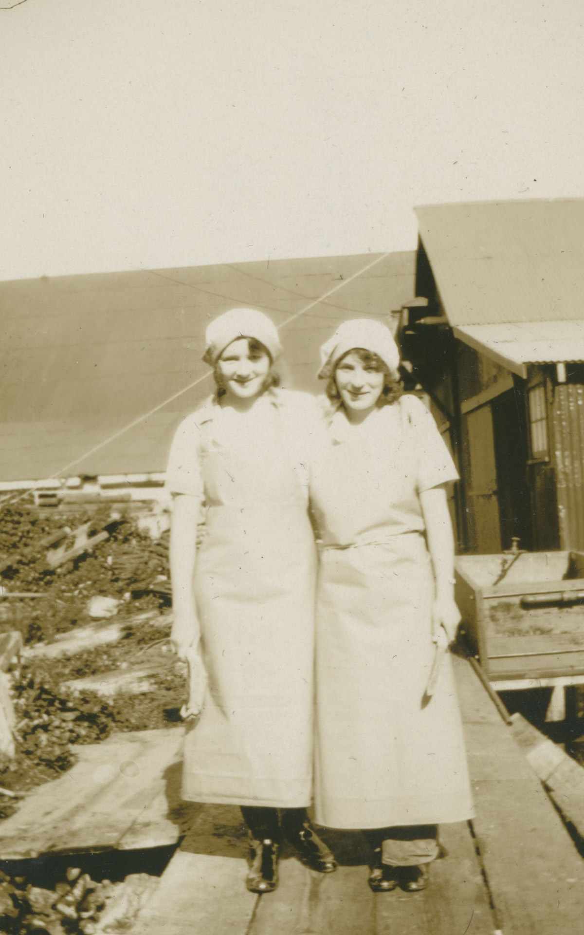 Two women in work uniforms pose in front of cannery buildings.