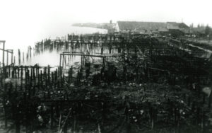 Piliers brûlés de plusieurs conserveries sur le bord de l'eau de Steveston avec la conserverie Gulf of Georgia intacte à l'arrière-plan.