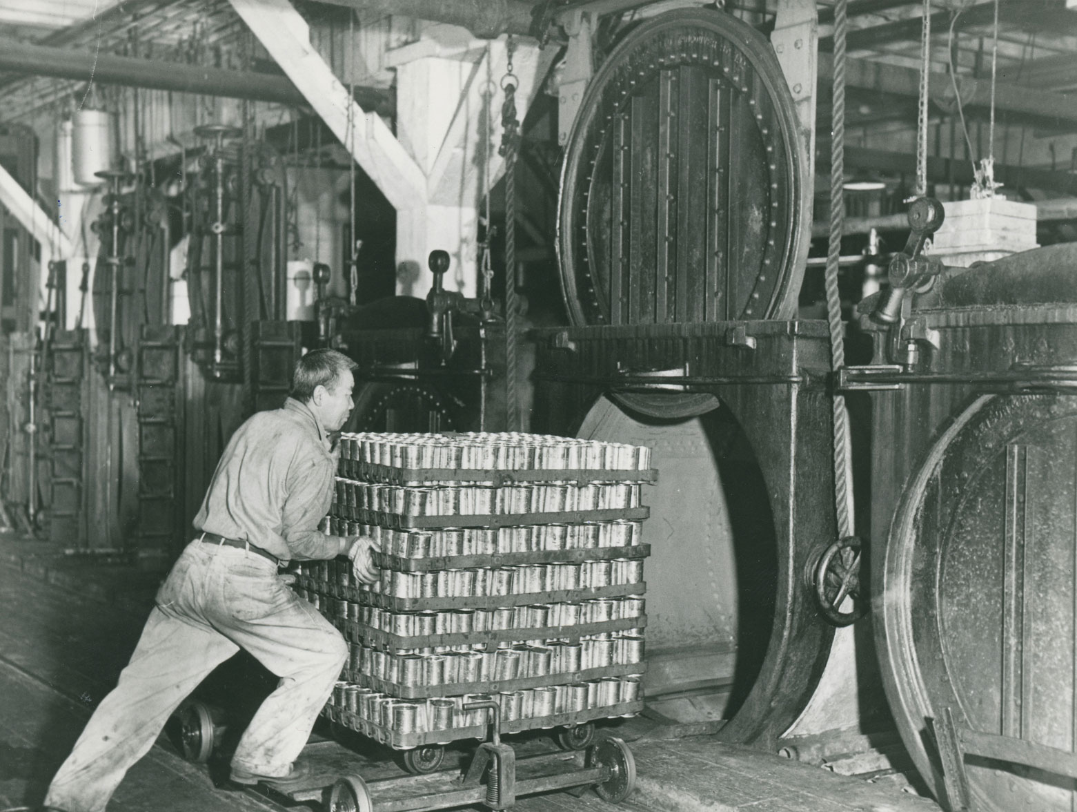 A man pushes a stack of seven trays of cans into an open retort oven. Four other ovens are in the background.