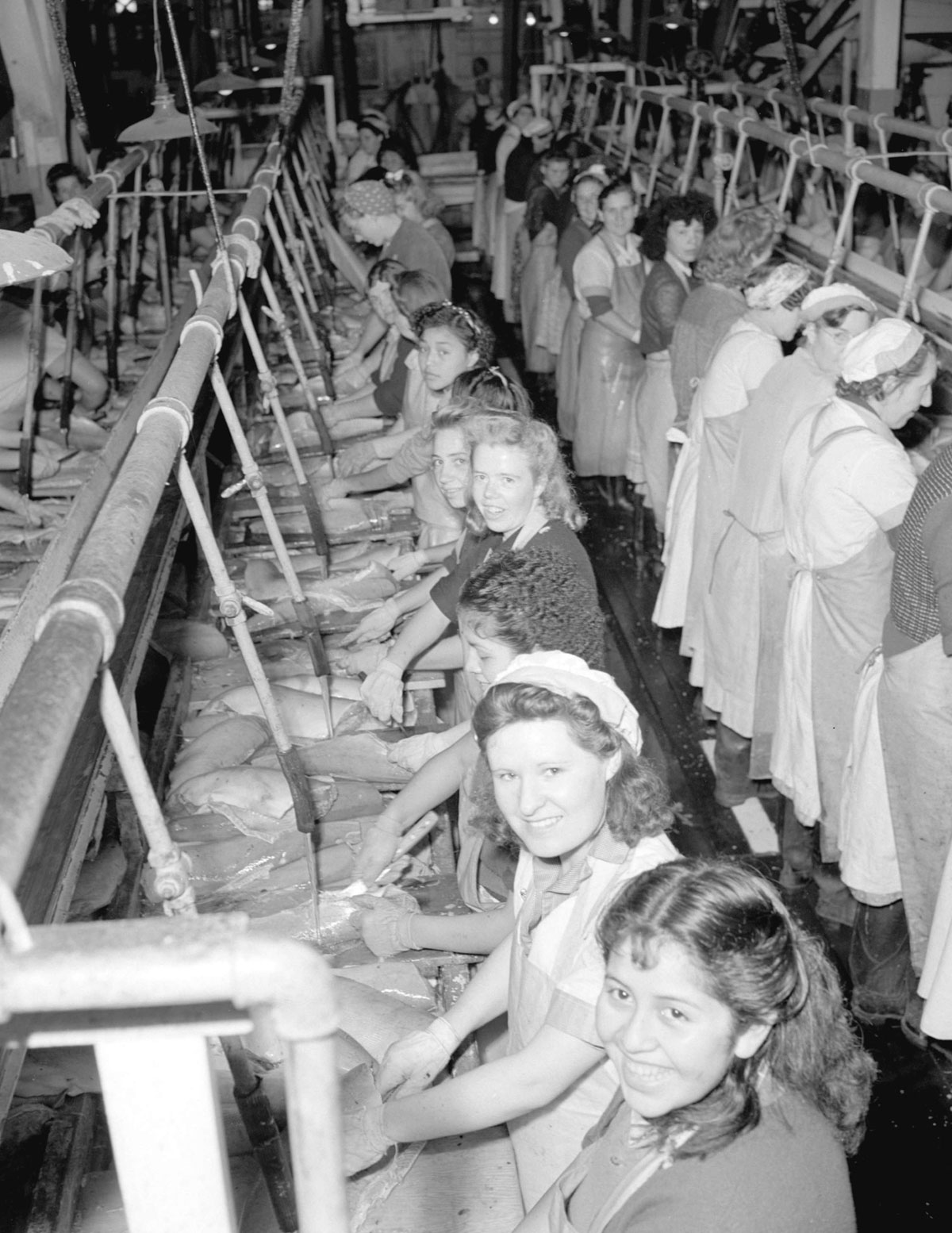 Women clean fish at cleaning tables. Several of the women are looking at the camera and smiling.