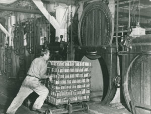 Un homme pousse une pile de sept plateaux de conserve dans un autoclave ouvert. Quatre autres fours se trouvent à l'arrière-plan.