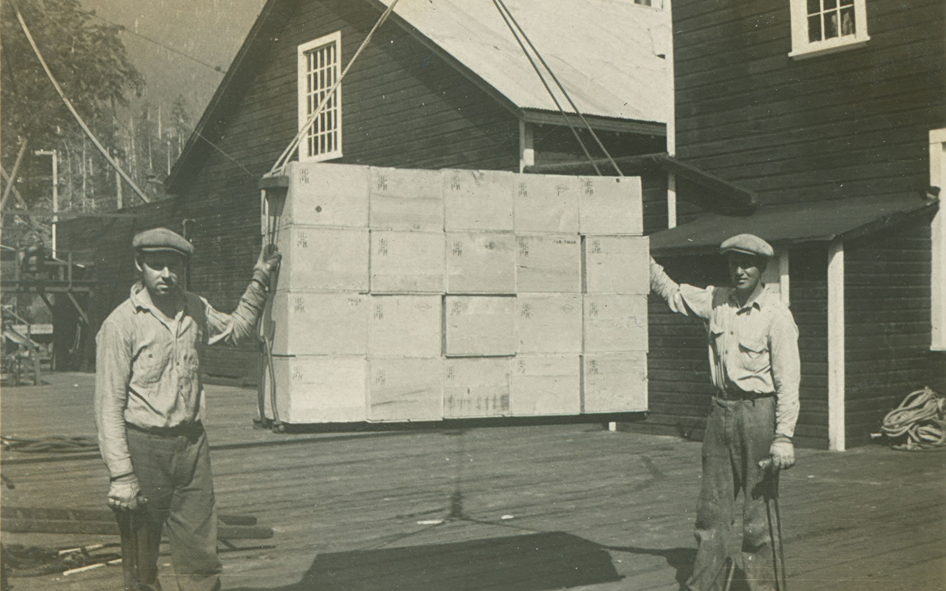 Two men stand on either side of stacked shipping crates suspended by pulley above a cannery dock.