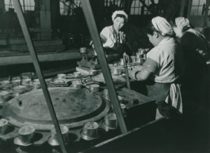 Trois ouvrières travaillent à une table à côté de laquelle se trouve une machine qui pèse les boîtes à l'aide d'un mécanisme circulaire.