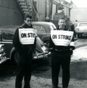 Deux hommes, devant des bâtiments et des voitures stationnées brandissent des écriteaux sur lesquels on peut lire « on strike » (en grève).