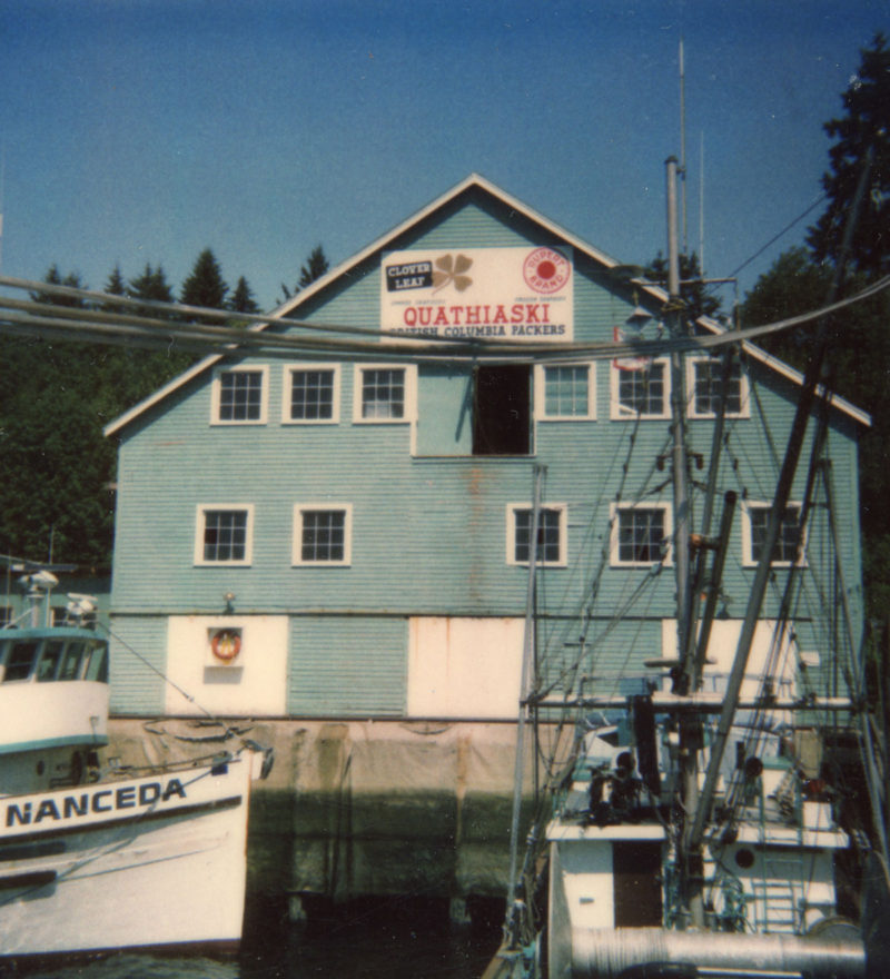 Bâtiment bleu avec deux rangées de grandes fenêtres donnant sur l'eau. Des bateaux de pêche à l'avant-plan