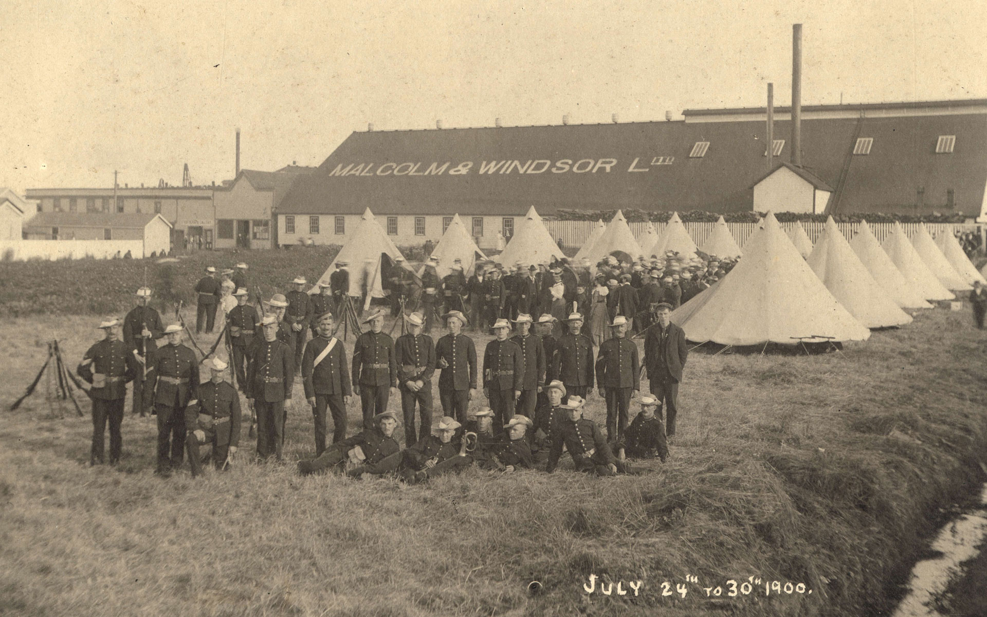 Soldiers stand together in front of tents on a grassy field. A building is in the background with Malcolm and Windsor Ltd. in large letters on the roof.
