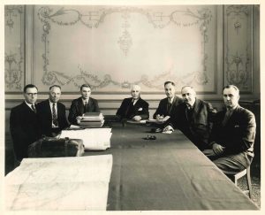 Photo en noir et blanc de sept hommes assis autour d’une longue table.