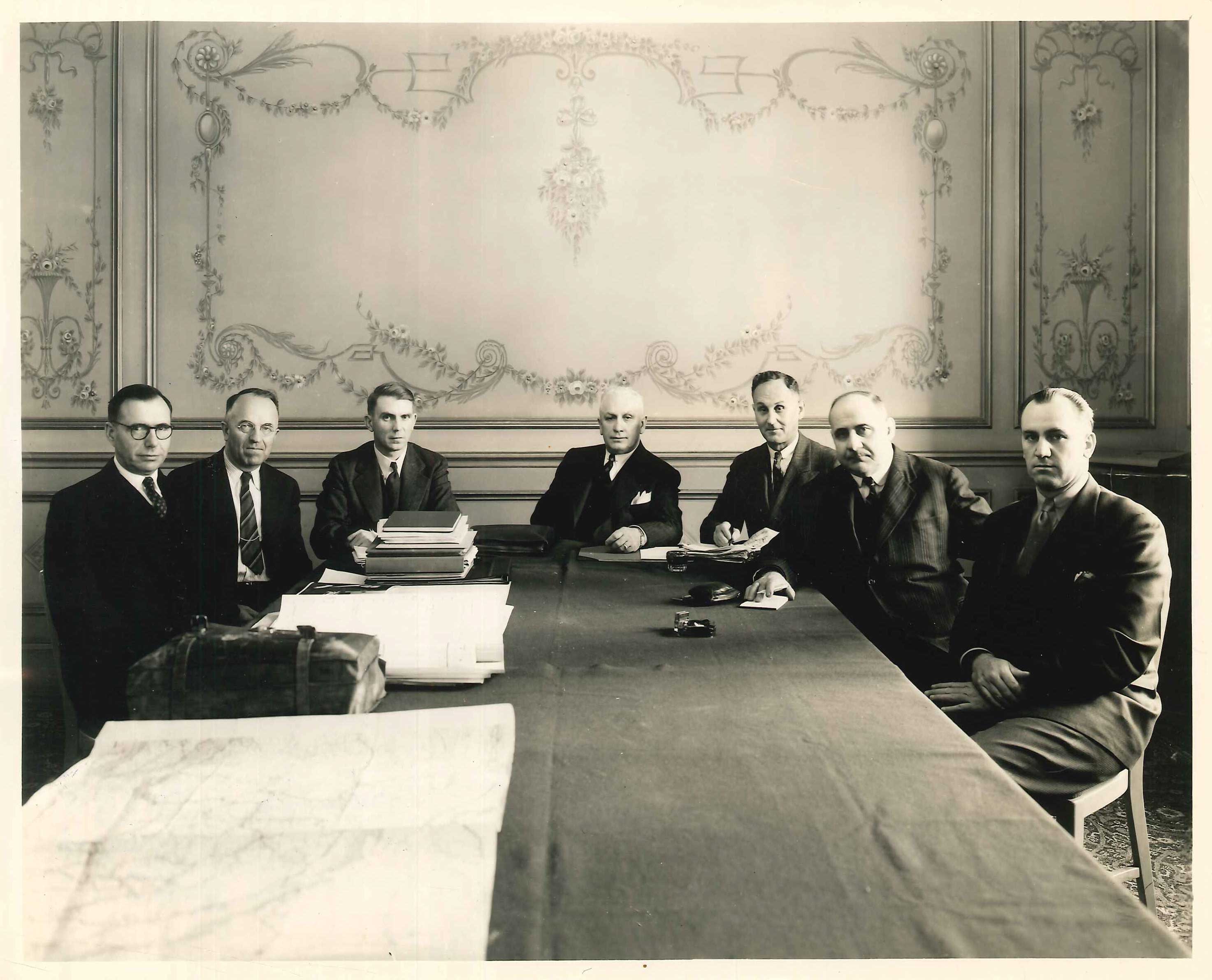 Black and white photograph of seven men sitting around a long table.