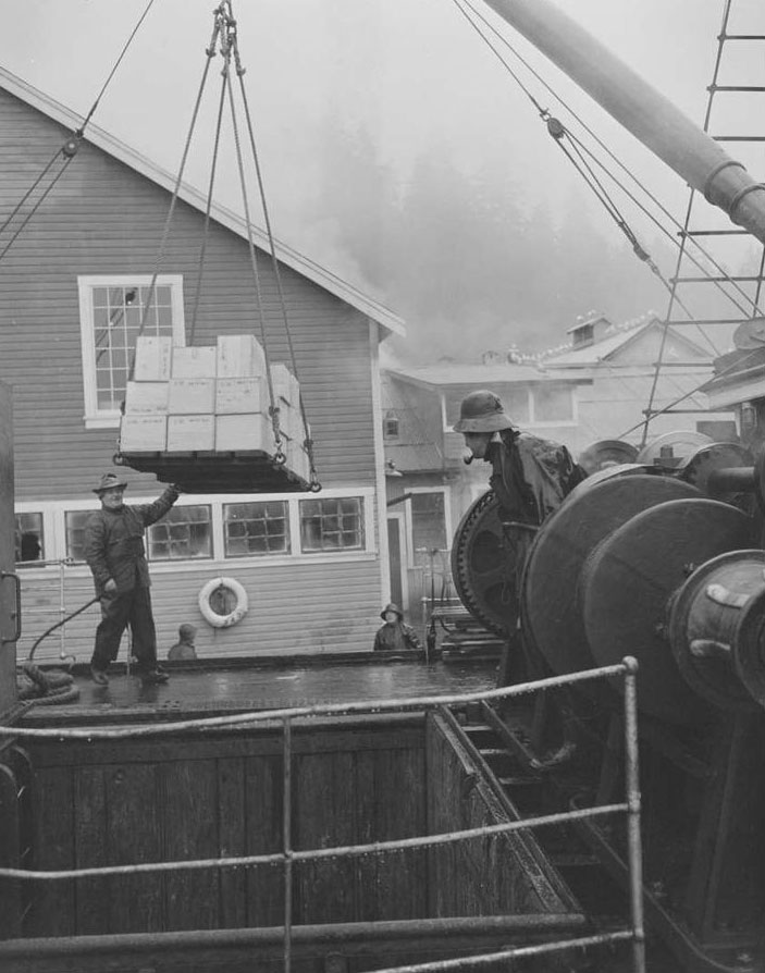 Workers look on as crates of canned salmon are loaded from the cannery dock into the hold of the boat with a large pulley.