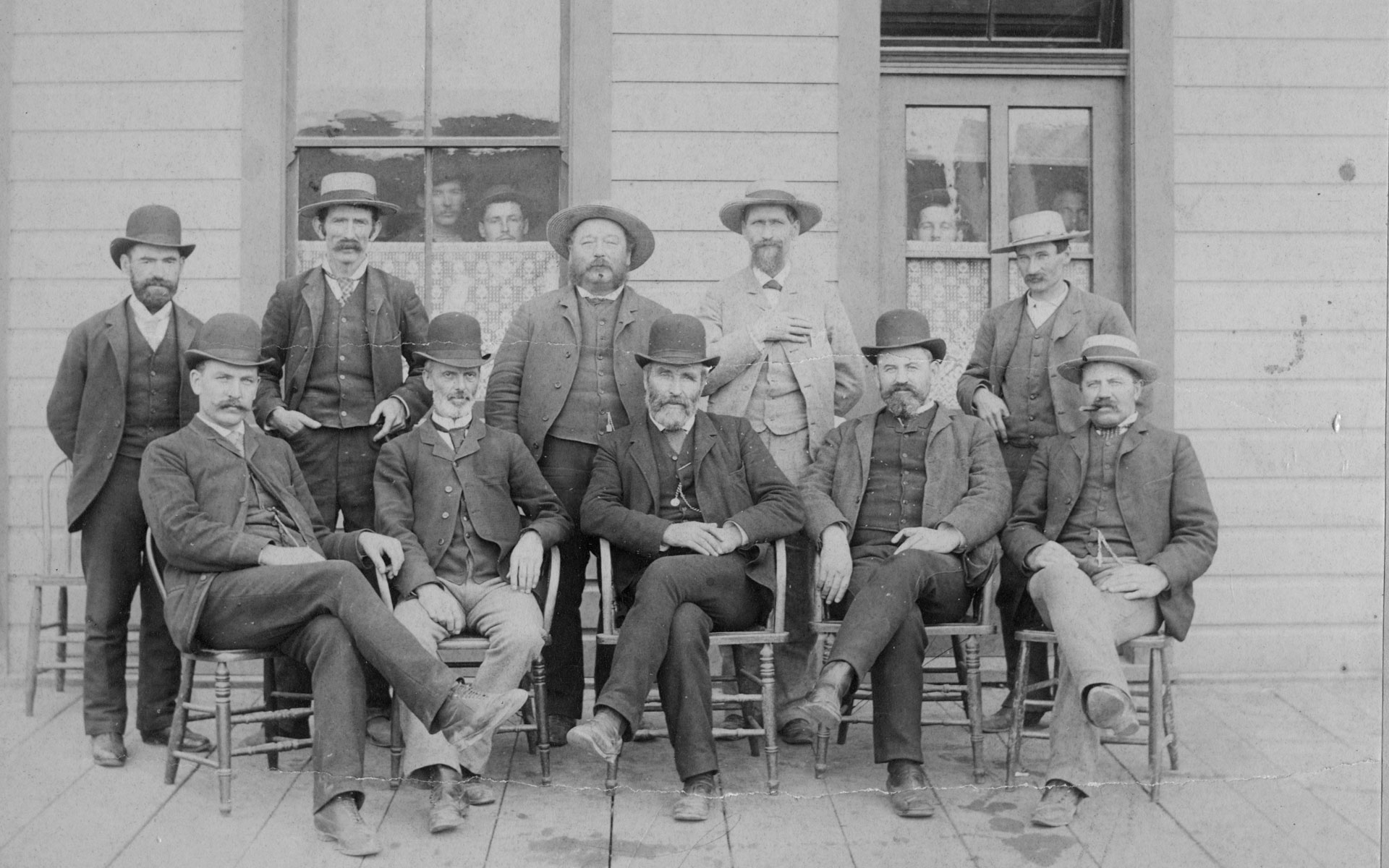 Group portrait with 5 men seated and 5 men standing behind. Four additional people are peaking out the windows of the building behind the men.
