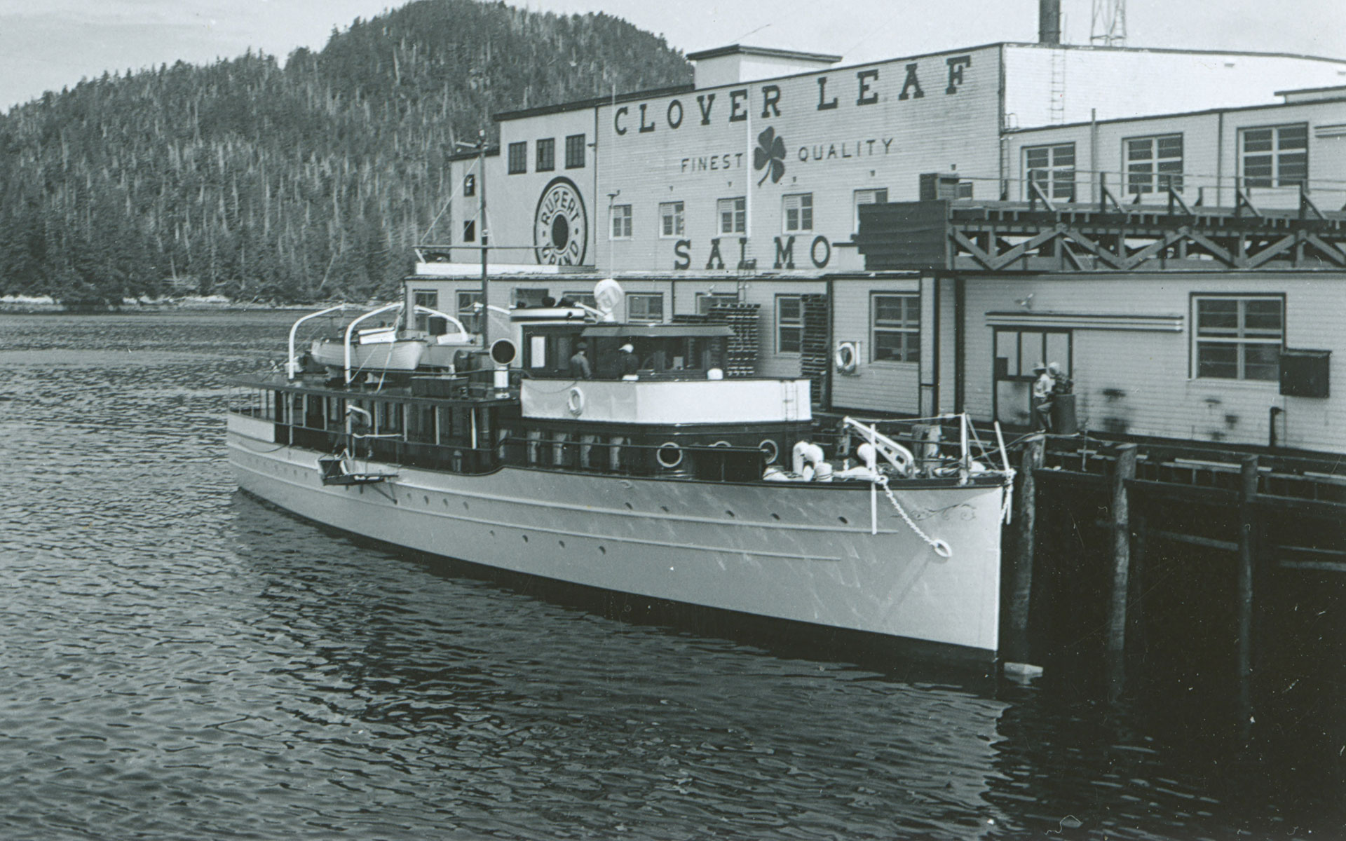 Passenger vessel docked at the Namu cannery. The words "Clover Leaf finest quality salmon" and "Rupert Brand" are painted on the side of the cannery building.