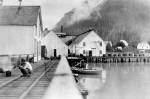 A man is squatting on the wharf with cannery buildings in the background.