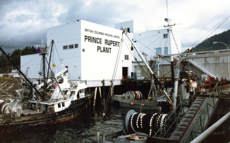 La conserverie Oceanside avec en avant-plan des quais et des bateaux de pêche. La mention « British Columbia Packers ltée Usine de Prince-Rupert » est peinte sur le bâtiment.