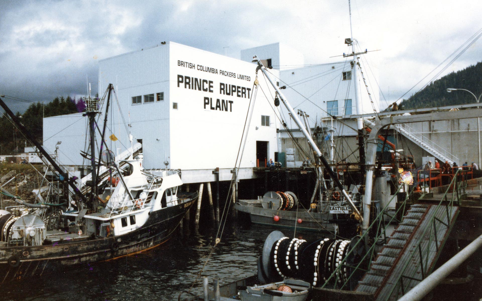 The Oceanside Cannery with docks and fishing boats in the foreground. The building is painted with the words "British Columbia Packers Limited Prince Rupert Plant."