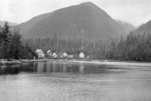 Cannery buildings nestled at the end of a bay with a treed mountain behind.