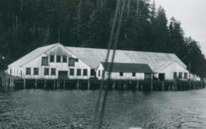Les bâtiments de la conserverie Shushartie photographiés d'un bateau sur l'eau. En avant-plan, deux cordes faisant partie de l'équipement du bateau. La conserverie est située au bord de l'eau devant une colline boisée.