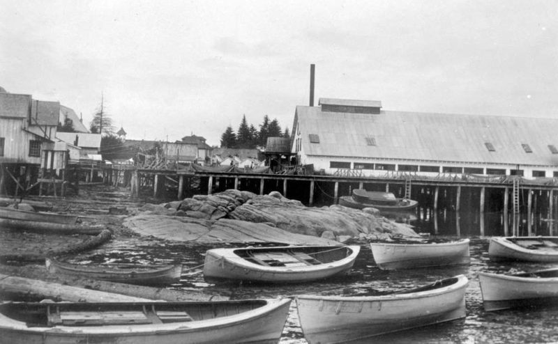 Skiffs are lined up along the beach at low tide in front of cannery buildings.