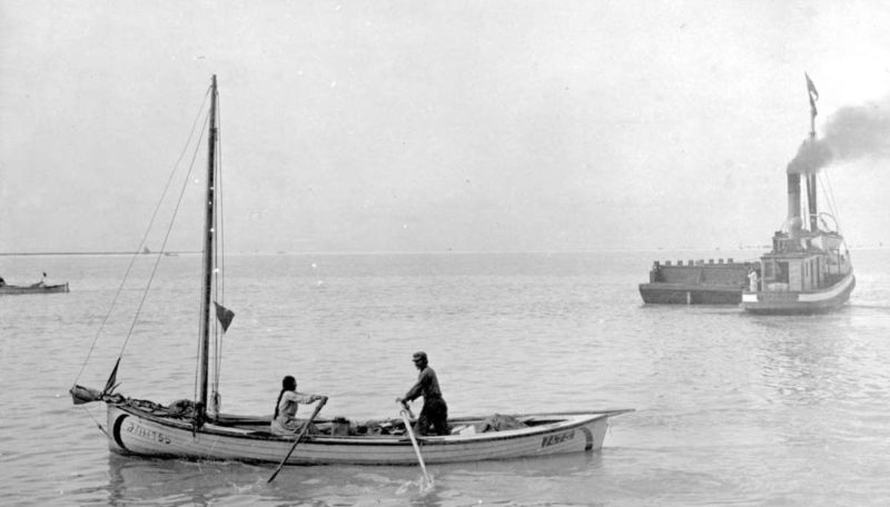 Des membres des Premières Nations pagaient sur un skiff sur le fleuve Fraser en 1913. Un bateau-remorque à vapeur se trouve en arrière-plan avec un chaland à ses côtés.