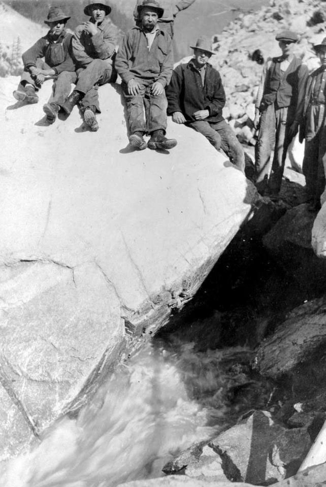 Travailleurs assis et debout aux côtés d'un immense rocher sur la rive du fleuve Fraser.