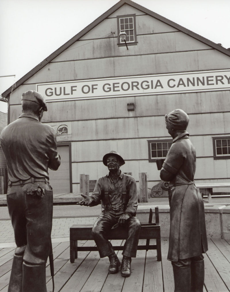 Statues d'un ramendeur assis sur un banc et de deux ouvriers debout devant lui. En arrière-plan, la conserverie Gulf of Georgia.