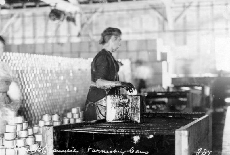 A worker stands at a table varnishing cans. Script at the bottom of the image reads: "B.C. Canneries. Varnishing Cans. FDT."