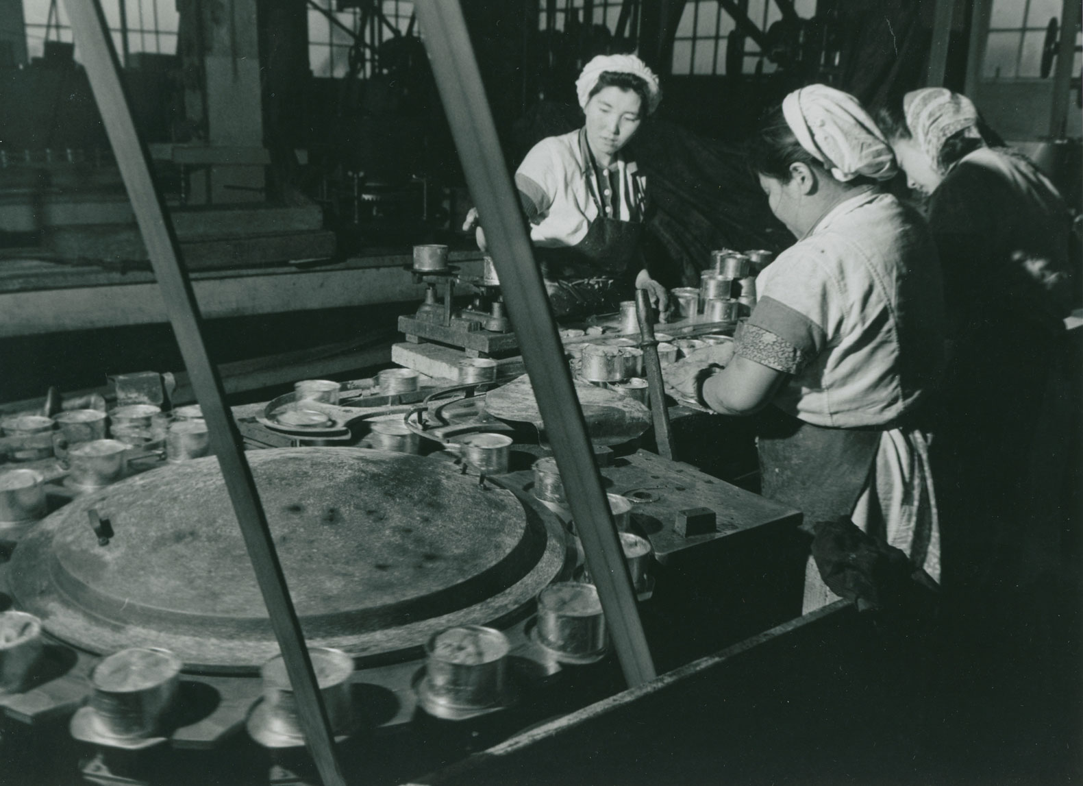 Trois ouvrières travaillent à une table à côté de laquelle se trouve une machine qui pèse les boîtes à l'aide d'un mécanisme circulaire.