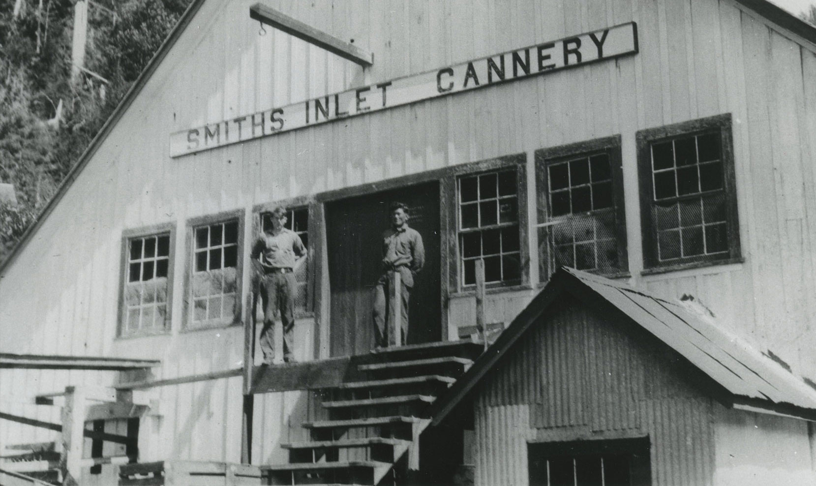 Two men stand at the top of steps leading into the wooden cannery building